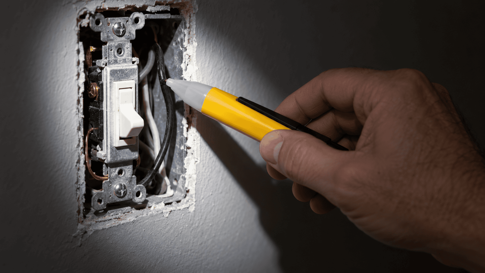 Hand holding a non contact voltage tester against the wires in an open light switch box
